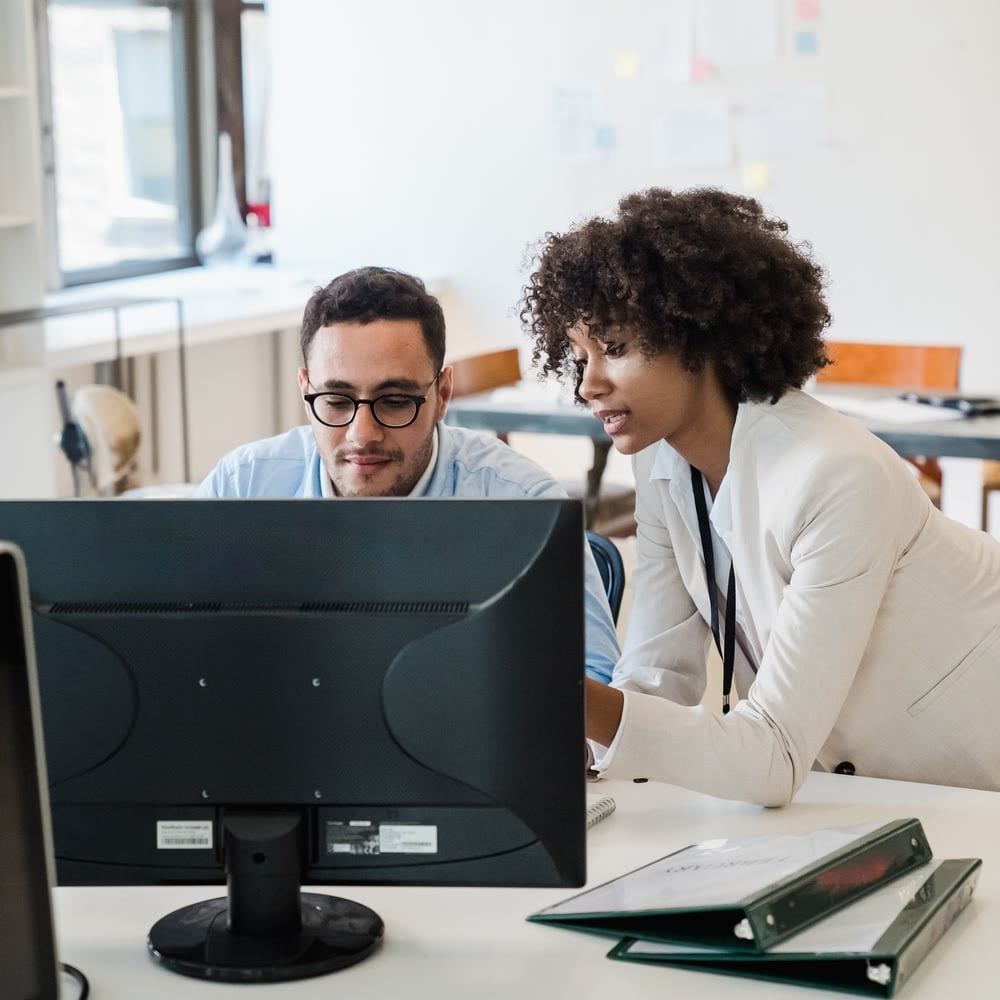 Two colleagues discussing something on a computer screen at a newsroom desk
